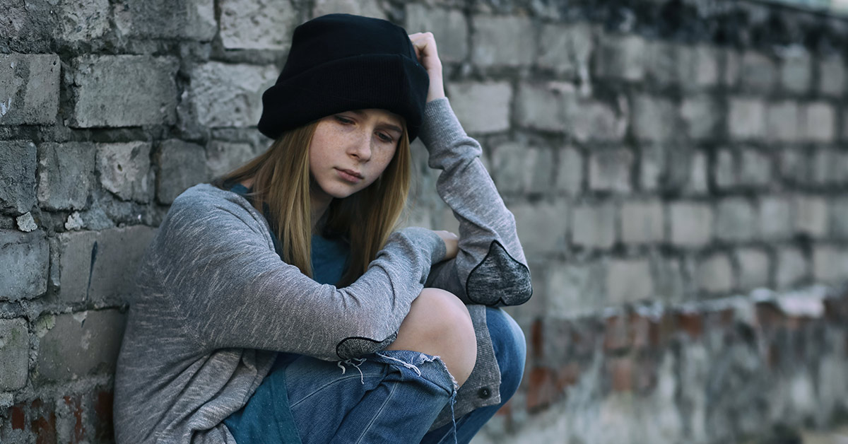girl sitting next to a wall wearing a black hat, grey shirt and ripped blue jeans